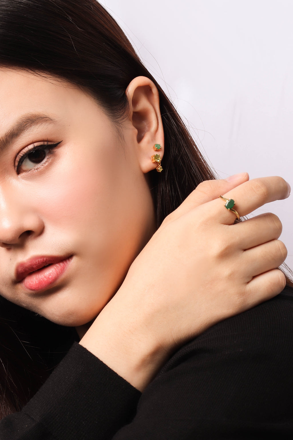 Close-up of a woman wearing a gold ring with a green stone on her finger and matching gold and green stud earrings, shown against a plain light background.