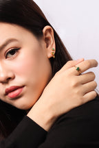 Close-up of a woman wearing a gold ring with a green stone on her finger and matching gold and green stud earrings, shown against a plain light background.