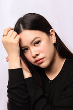 A close-up portrait of a young woman with long dark hair, wearing a black long-sleeve top and small gold earrings, posing with her hand on her head against a light gray background.