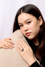 A woman with long dark hair wears a ring with a large oval blue gemstone and a gold band, resting her chin on a textured beige pillow against a light background.
