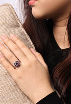 A close-up lifestyle shot of a woman wearing a silver ring with a dark purple and red gemstone cluster, resting her hand on a beige woven blanket.
