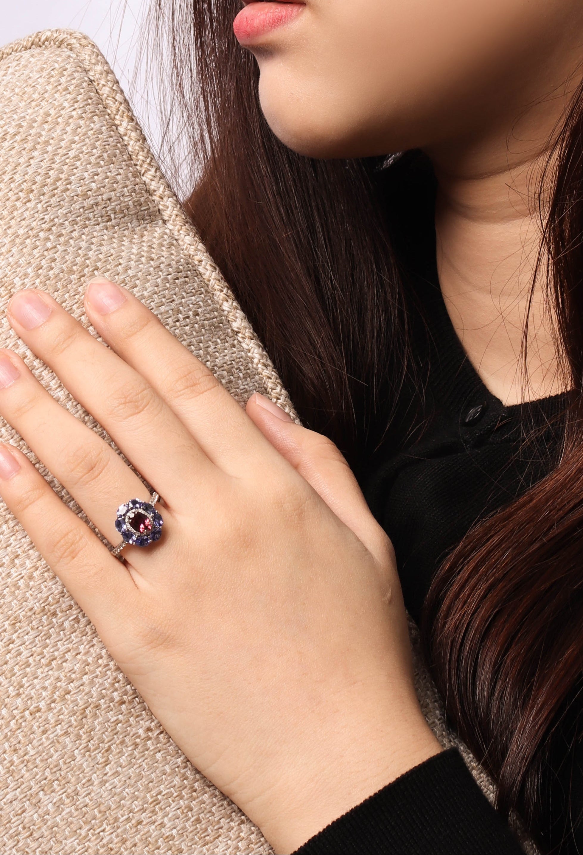 A close-up lifestyle shot of a woman wearing a silver ring with a dark purple and red gemstone cluster, resting her hand on a beige woven blanket.