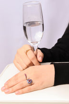A close-up of a person's hands resting on a white surface, with one hand wearing a silver-toned ring featuring a red heart-shaped gemstone and a purple chain detail. The other hand holds a clear glass of water.