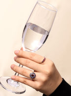 A hand holding a clear wine glass against a light beige background, wearing a silver ring with a dark purple and blue floral center and a smaller matching ring on the other finger.