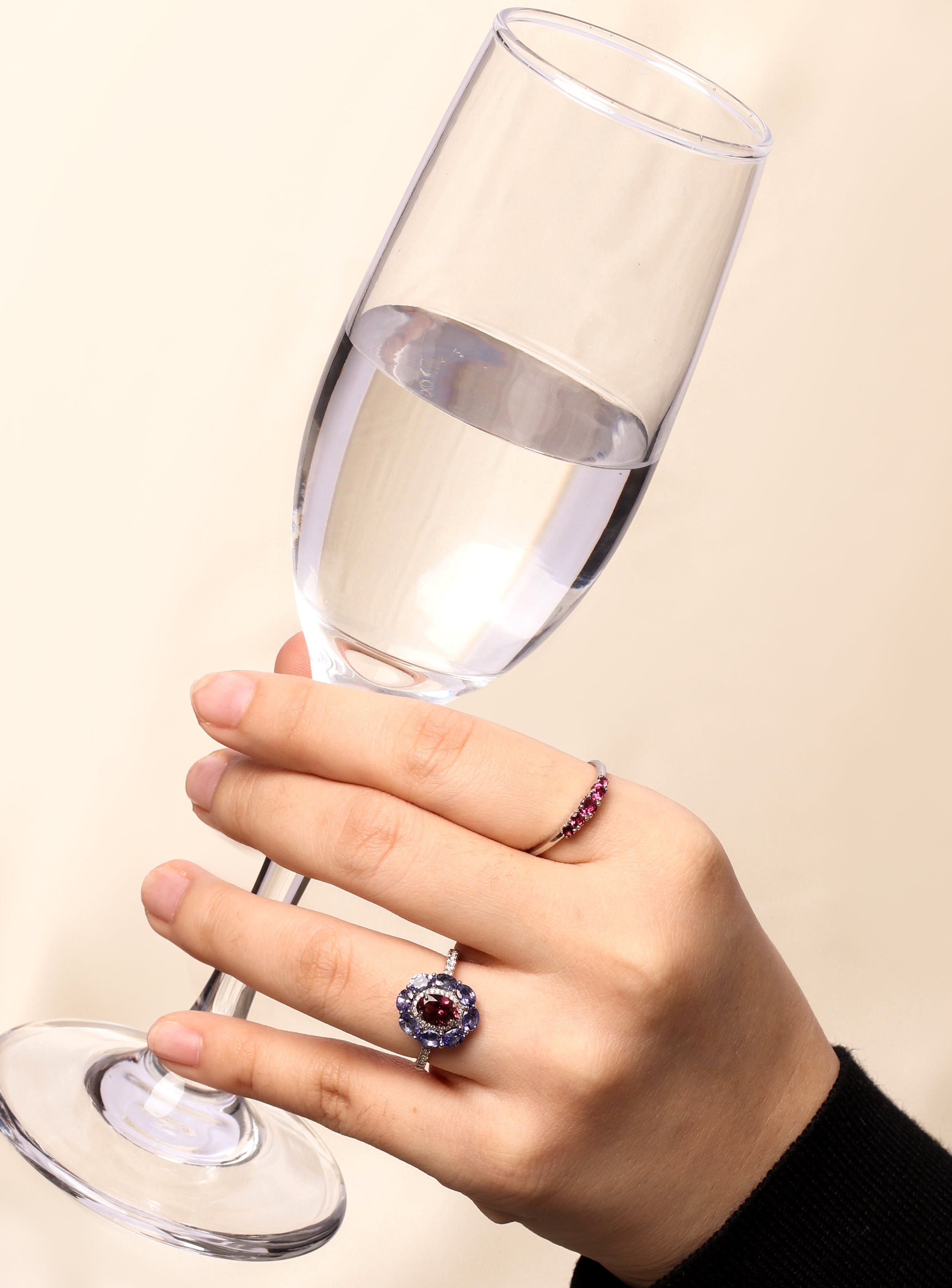 A hand holding a clear wine glass against a light beige background, wearing a silver ring with a dark purple and blue floral center and a smaller matching ring on the other finger.