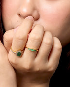 Close-up of a person wearing multiple rings on their fingers, including a prominent pink and gold ring, a green stone ring, and slim gold rings with green accents, against a light background.