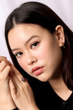A close-up portrait of a woman with dark hair wearing a small gold hoop earring and a ring with a red heart-shaped stone on a black top, against a light gray background.