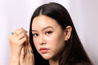 A close-up portrait of a woman with long dark hair, wearing a green gemstone ring on one finger and a small gold hoop earring, set against a plain light gray background.