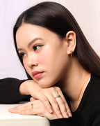 A close-up portrait of a woman with long dark hair resting her chin on her hands, wearing gold rings and a small gold earring. 