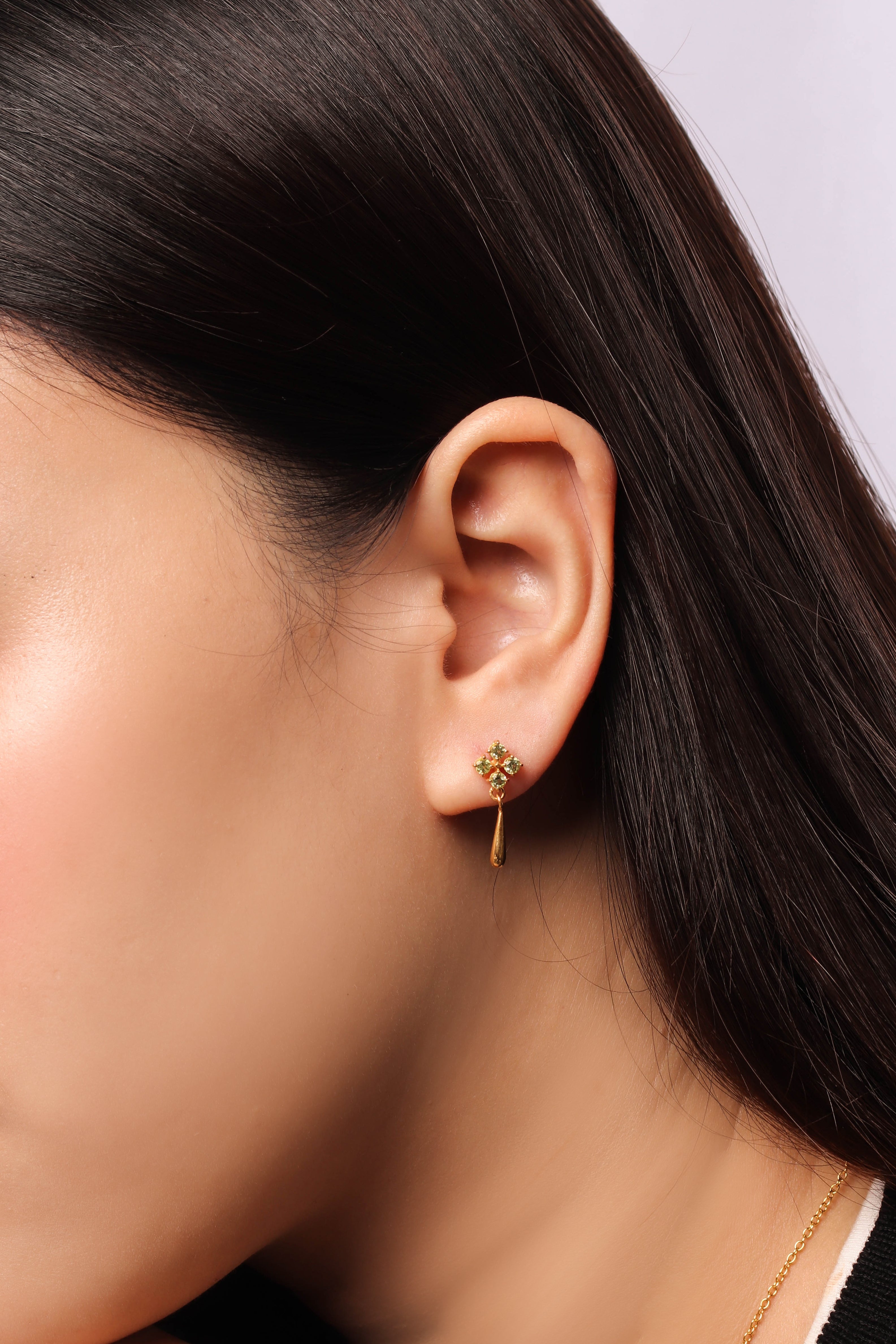 A close-up of a model's ear wearing a delicate gold dangle earring with a small floral cluster at the top.