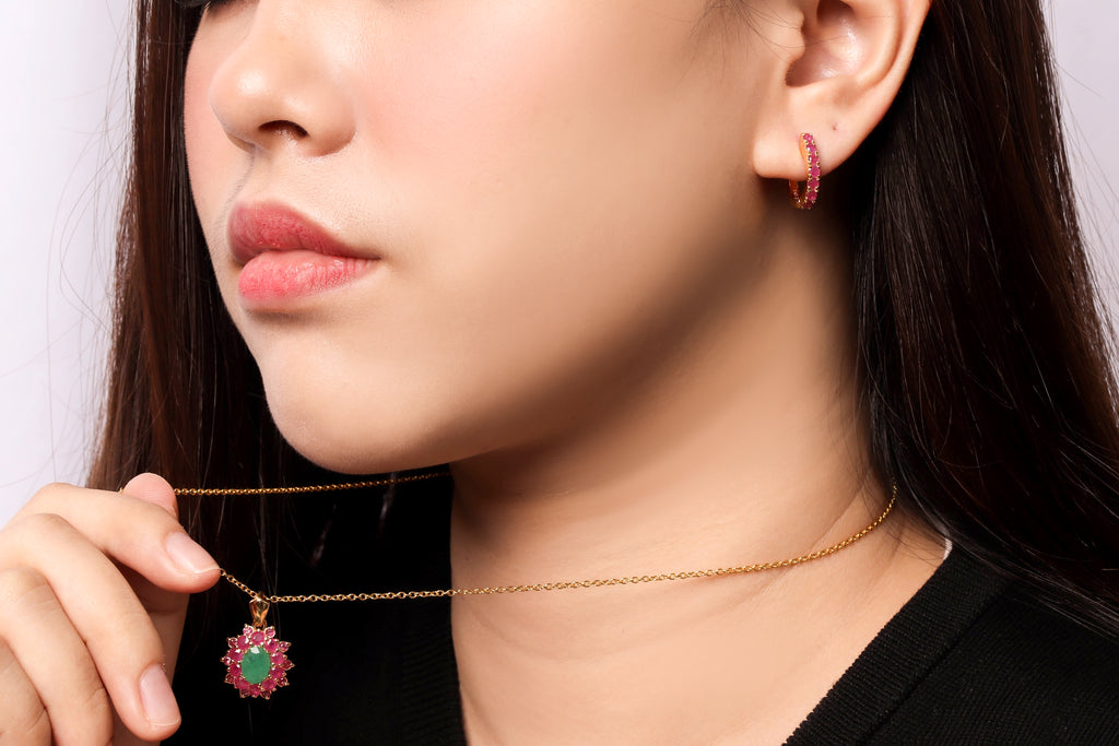 Close-up of a woman wearing a delicate gold chain necklace with a green heart-shaped pendant accented by pink stones, and matching pink hoop earrings.