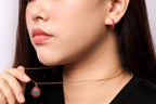 Close-up of a woman wearing a delicate gold chain necklace with a green heart-shaped pendant accented by pink stones, and matching pink hoop earrings.