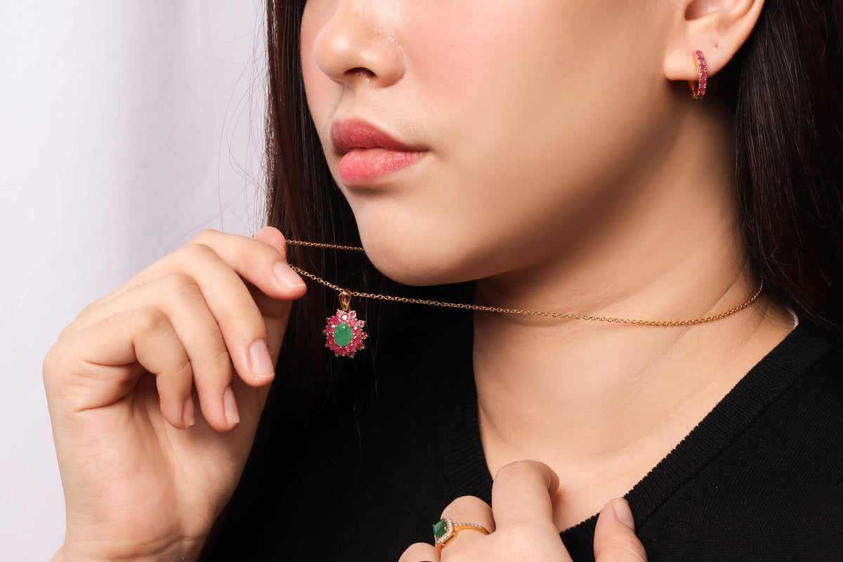 A close-up lifestyle shot of a model wearing a delicate gold chain necklace with a small, vibrant flower-shaped pendant featuring green and pink stones. The model is wearing a black top and has a matching gold hoop earring.