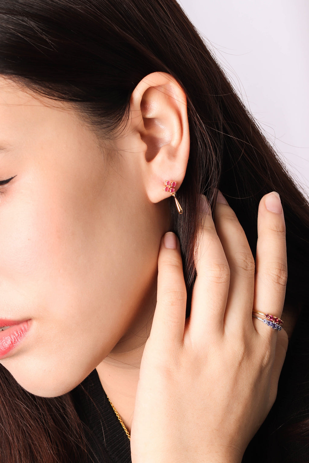 A close-up lifestyle shot of a woman wearing a delicate rose gold huggie hoop earring with a small red crystal accent, along with a matching rose gold ring on her finger.
