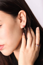 A close-up lifestyle shot of a woman wearing a delicate rose gold huggie hoop earring with a small red crystal accent, along with a matching rose gold ring on her finger.