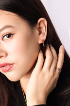 Close-up of a woman wearing a delicate gold dangle earring with a small red gemstone and a matching gold ring with red and blue stones.