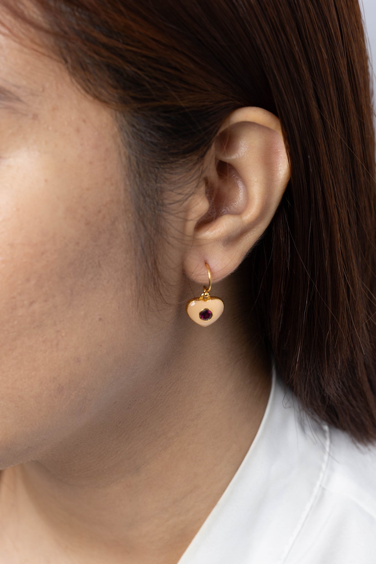 A close-up of a person's ear wearing a small gold heart-shaped dangle earring with a dark red center stone.