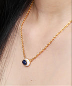 A close-up of a gold chain necklace worn on a model's neck, featuring a small circular pendant with a dark blue center and a white outer ring.