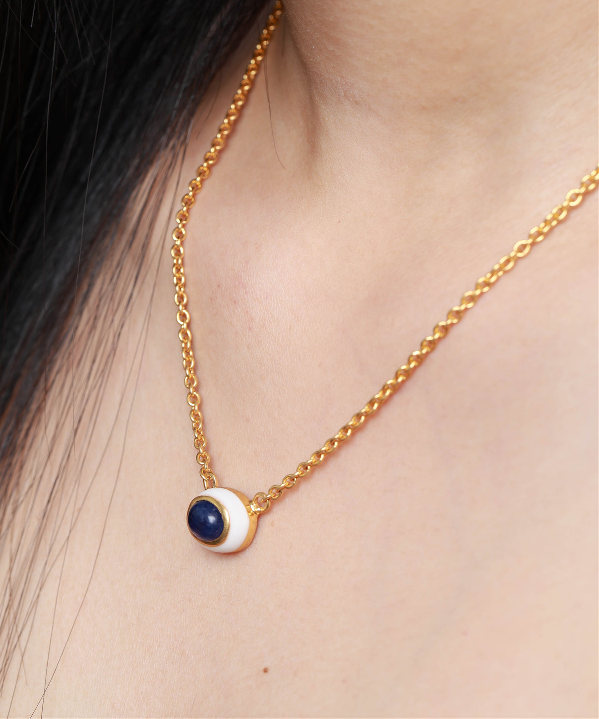 A close-up of a gold chain necklace worn on a model's neck, featuring a small circular pendant with a dark blue center and a white outer ring.