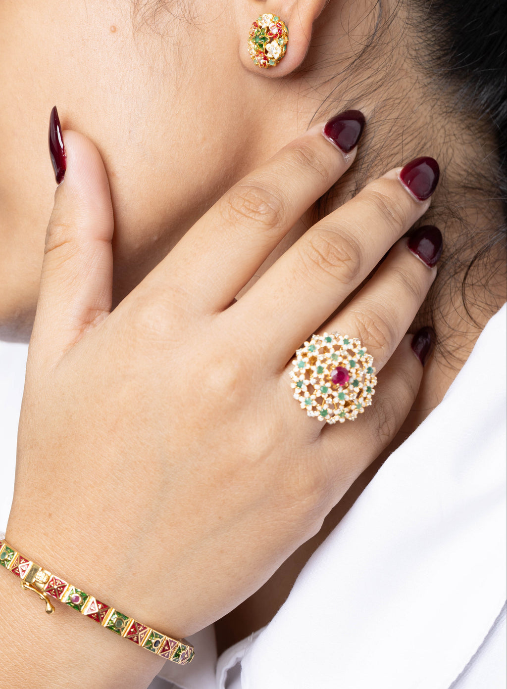A close-up lifestyle shot of a woman wearing a gold-plated Kundan jewelry set, including a ring with a central red stone, matching stud earring, and bracelet, with her hand resting near her neck.