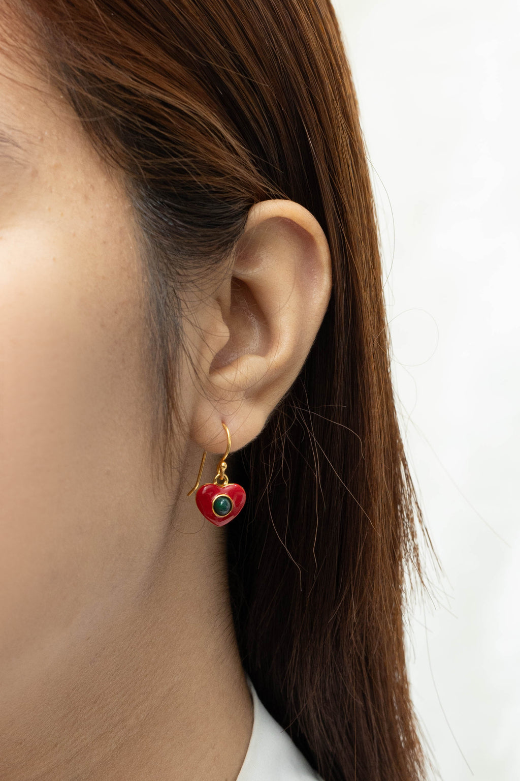 A close-up of a person's ear wearing a small dangling heart-shaped earring with red enamel, gold-tone metal, and a green circular detail.