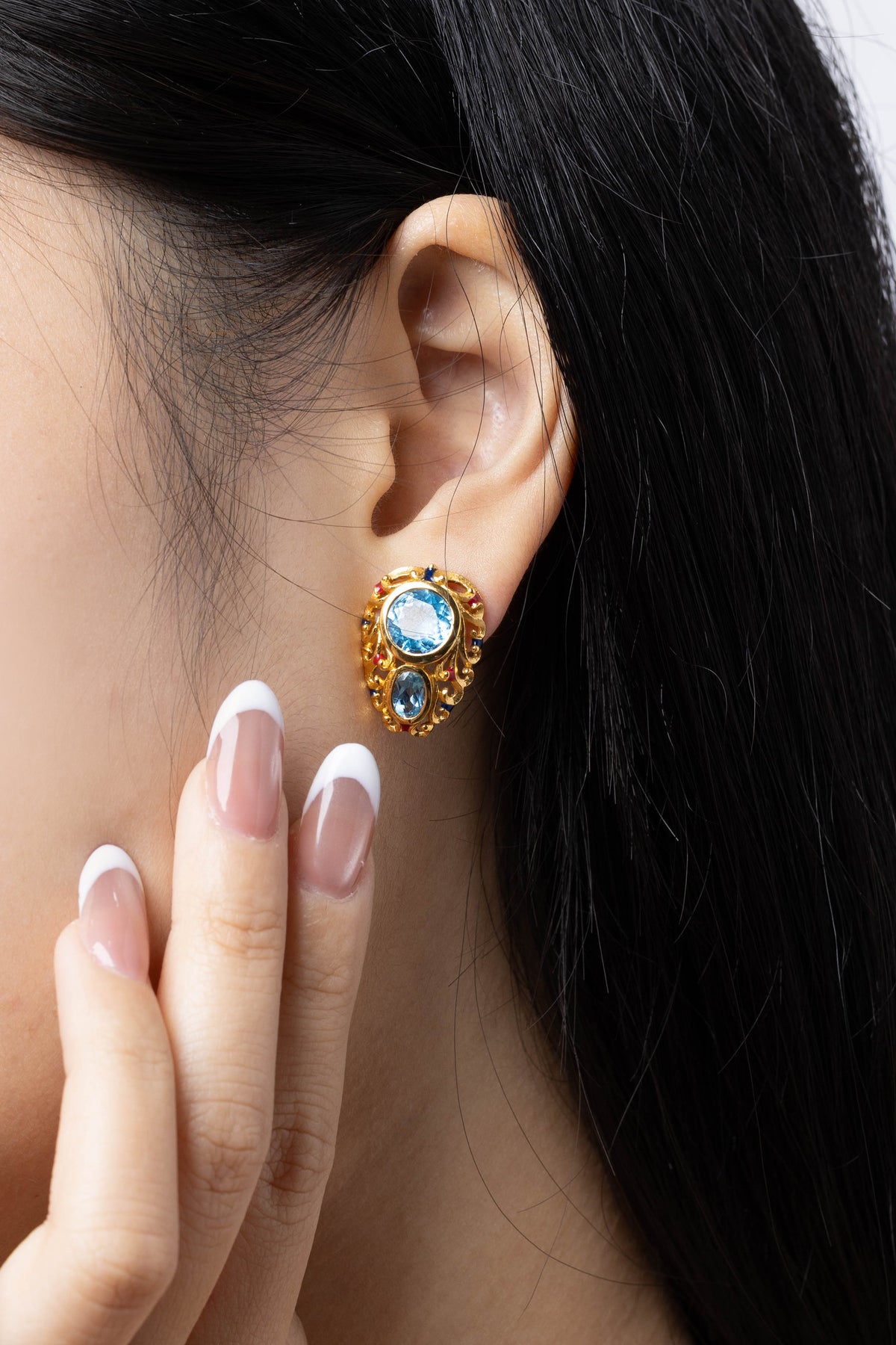 A close-up lifestyle shot of a model wearing a gold-plated earring with a light blue oval center stone and smaller blue stones, with her hand resting near her face.