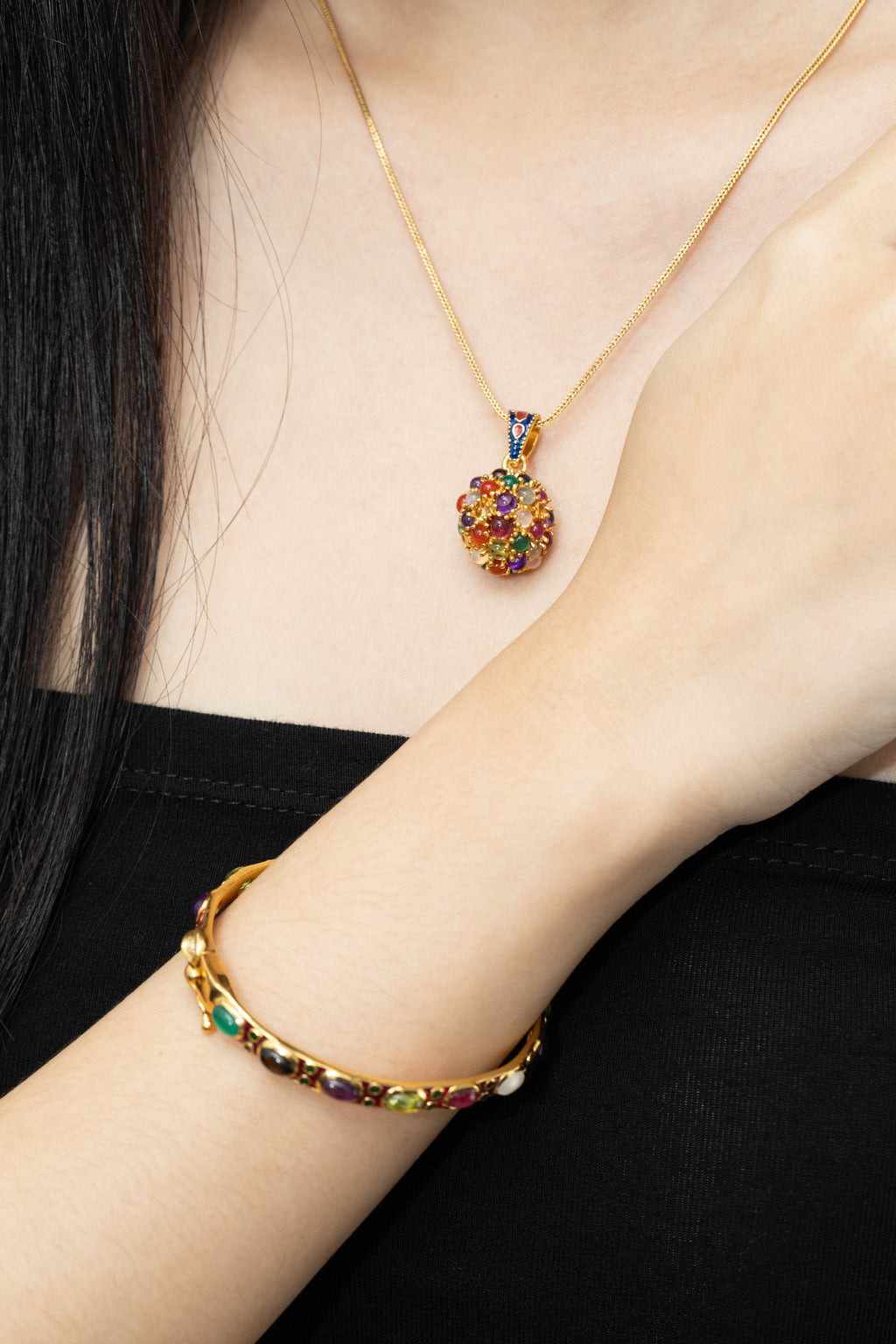 A close-up of a person wearing a gold necklace with a colorful gemstone pendant and a matching gold bracelet on the wrist, against a black top background.