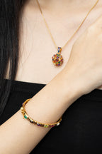 A close-up of a person wearing a gold necklace with a colorful gemstone pendant and a matching gold bracelet on the wrist, against a black top background.