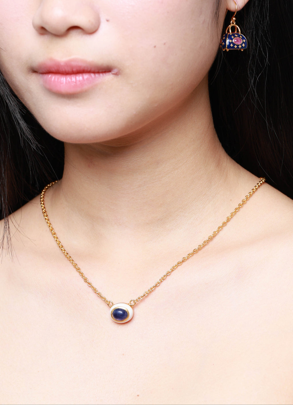A close-up of a model wearing a delicate gold chain necklace with a small oval pendant featuring a dark blue center and a gold border, along with a matching gold and blue stud earring.