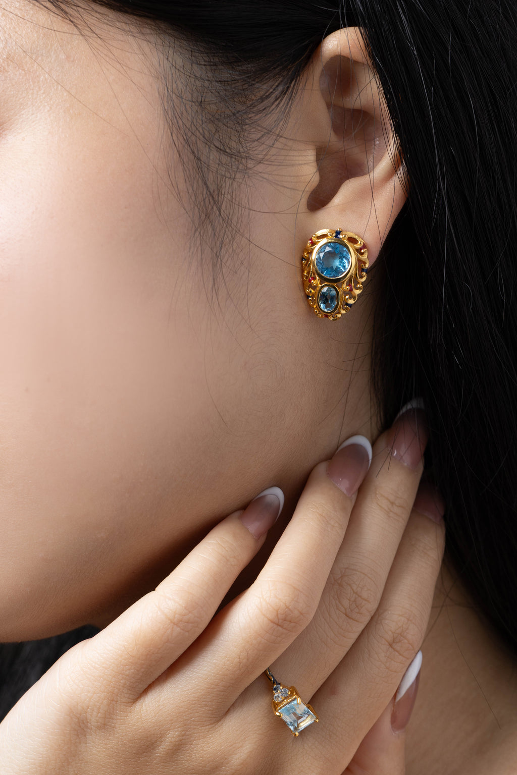 A close-up of a model's ear wearing a gold-toned stud earring with blue gemstones, alongside a matching gold ring on her finger.