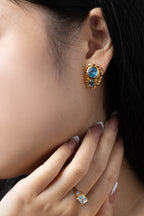 A close-up of a model's ear wearing a gold-toned stud earring with blue gemstones, alongside a matching gold ring on her finger.