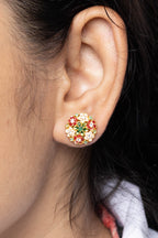 A close-up of a person's ear wearing a small floral stud earring with yellow-gold metal and red, green, and cream rhinestones.