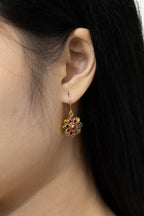 A close-up of a woman's ear wearing a gold-toned flower-shaped earring with multicolored enamel details in red, green, and blue.