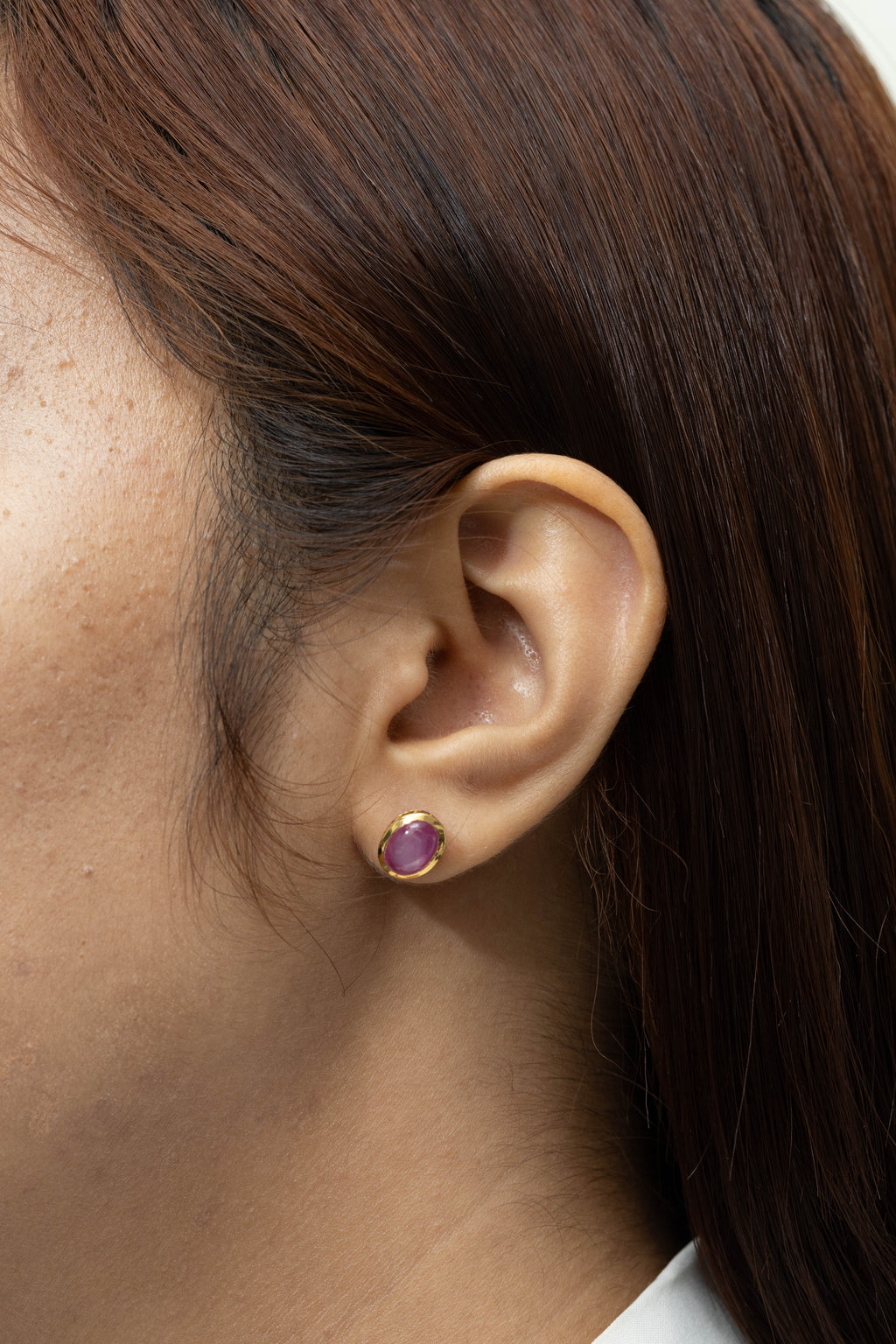 A close-up of a person's ear wearing a small stud earring with a round purple gemstone in a gold setting, against dark brown hair.