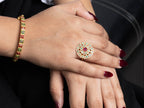 A close-up of a person's hands showcasing jewelry, featuring a large gold ring with a red center stone and surrounding gold and white stones, alongside a gold bracelet with red and green stones.