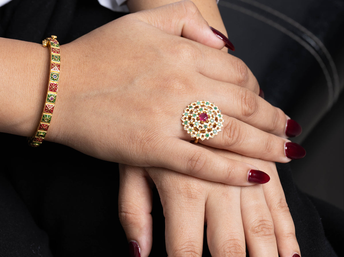 A close-up of a person's hands showcasing jewelry, featuring a large gold ring with a red center stone and surrounding gold and white stones, alongside a gold bracelet with red and green stones.
