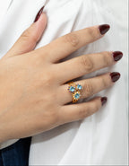 A close-up of a hand with dark burgundy nail polish resting on white fabric, wearing a gold ring with three light blue gemstones.