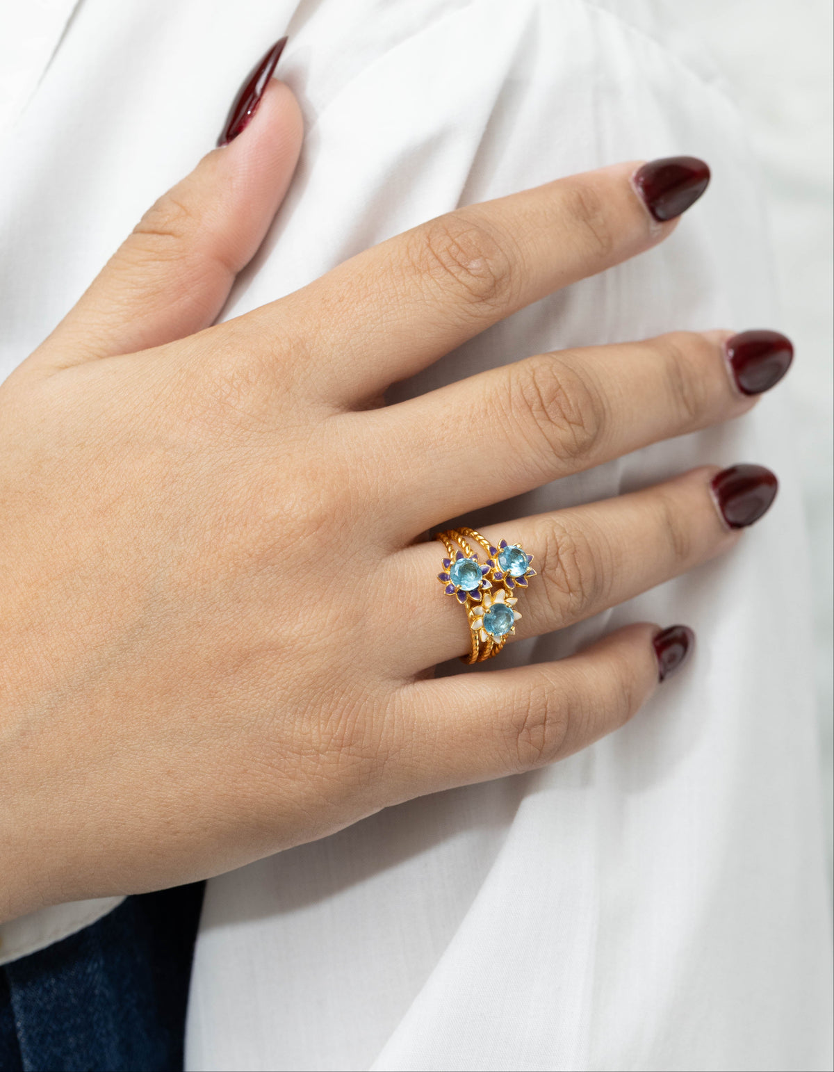 A close-up of a hand with dark burgundy nail polish resting on white fabric, wearing a gold ring with three light blue gemstones.
