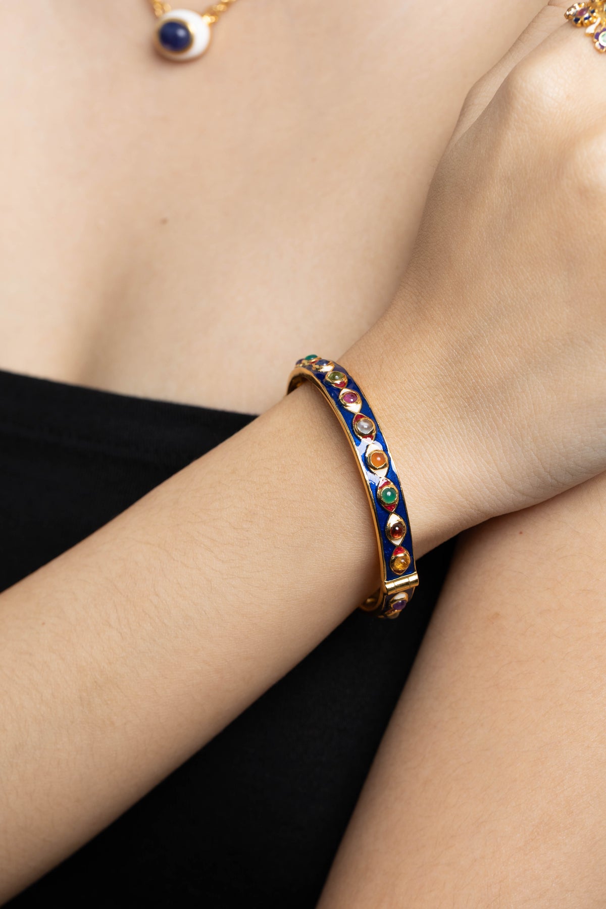A gold-toned bangle bracelet with a dark blue enamel center and colorful circular accents, worn on a person's wrist against a black strap and skin background.