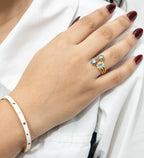 Gold ring with blue topaz gemstones and enamel flowers, worn on hand with white enamel bracelet