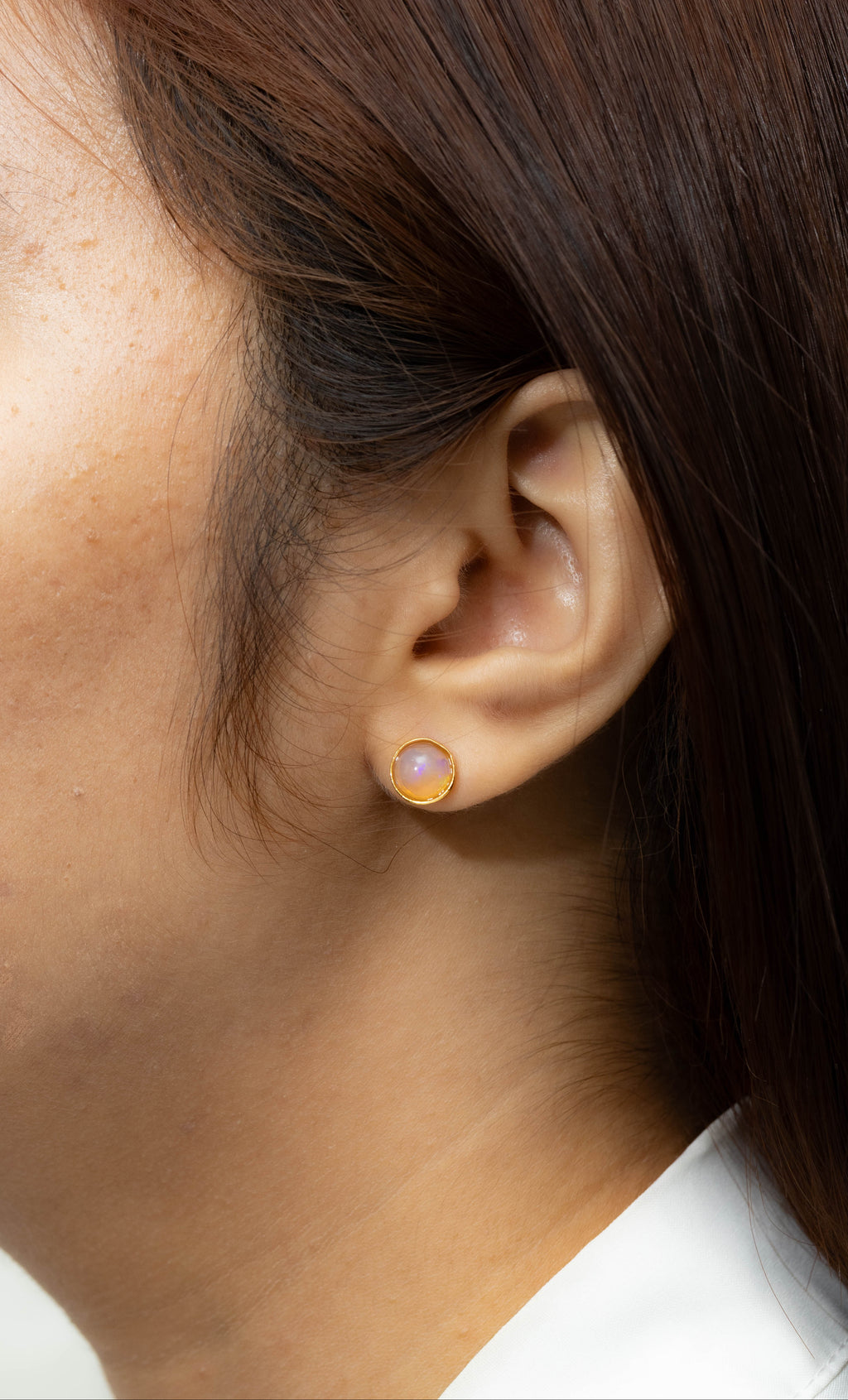A close-up side profile of a woman's ear wearing a small gold stud earring with a round iridescent lavender-pink gemstone.