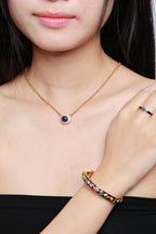 A close-up lifestyle shot of a model wearing a gold necklace with a dark blue oval pendant, a matching gold ring with colorful stones, and a gold bangle with colorful stones.