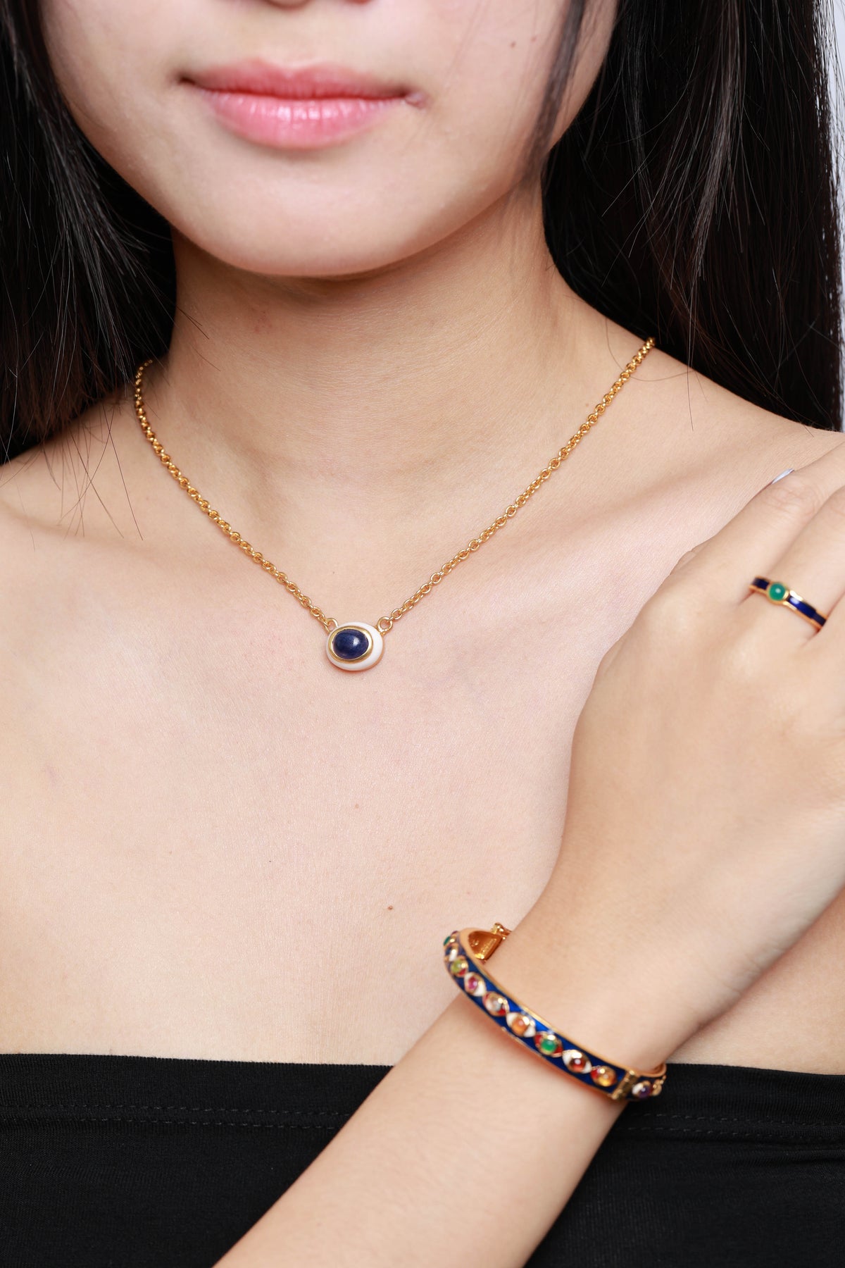 A close-up lifestyle shot of a model wearing a gold necklace with a dark blue oval pendant, a matching gold ring with colorful stones, and a gold bangle with colorful stones.
