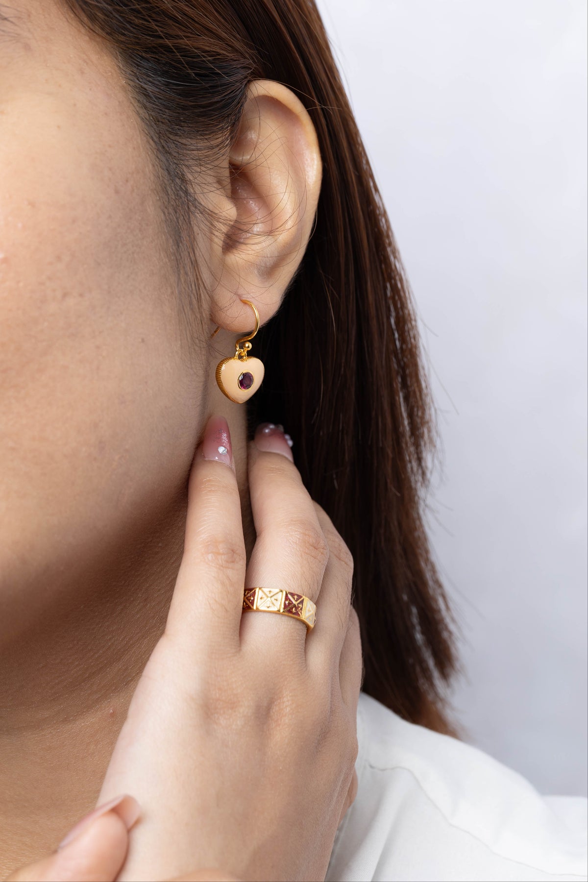 A close-up of a woman wearing a small gold heart-shaped drop earring with a dark red heart in the center, paired with a gold ring on her finger.