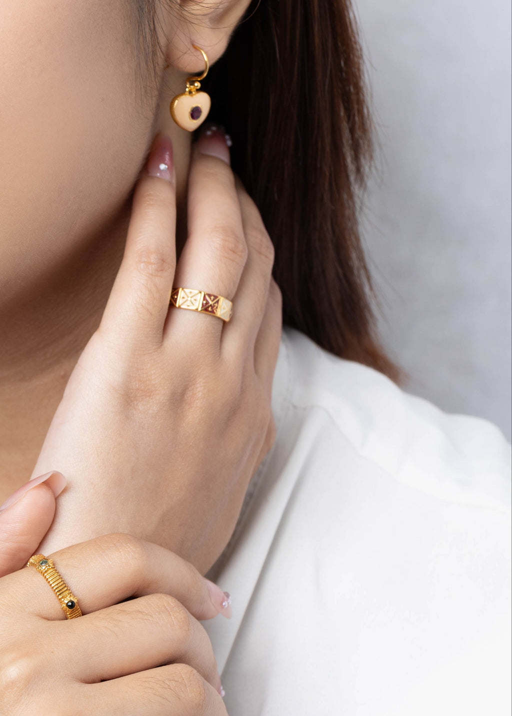 A close-up lifestyle shot of a woman wearing gold jewelry, featuring a small gold heart-shaped drop earring and gold rings on her fingers, set against a soft gray background.