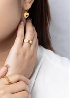 A close-up lifestyle shot of a woman wearing gold jewelry, featuring a small gold heart-shaped drop earring and gold rings on her fingers, set against a soft gray background.