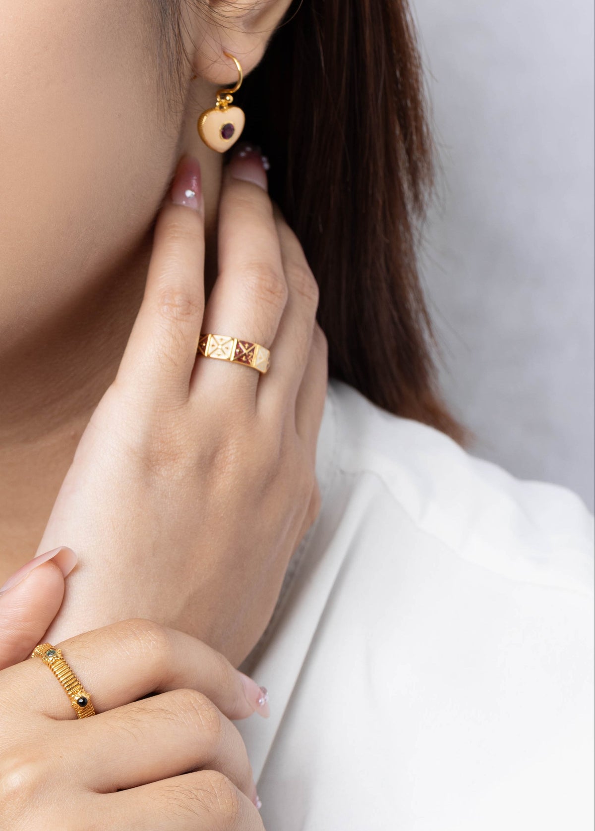 A close-up lifestyle shot of a woman wearing gold jewelry, featuring a small gold heart-shaped drop earring and gold rings on her fingers, set against a soft gray background.