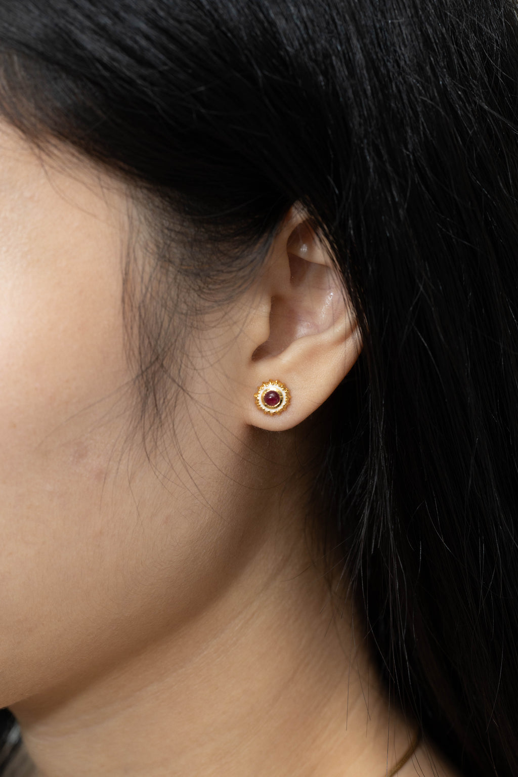 A close-up side profile of a person's ear wearing a small gold stud earring with a circular red gemstone in the center.