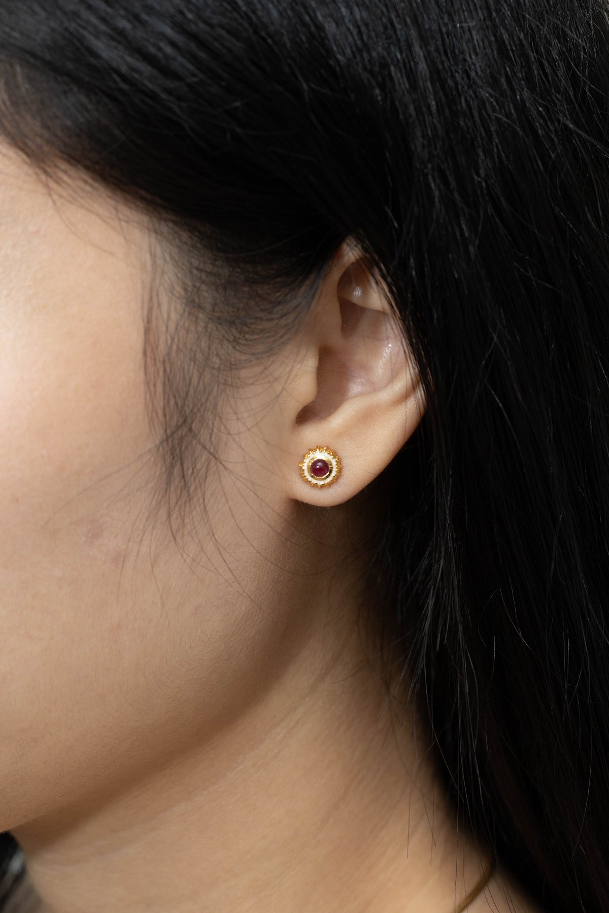 A close-up side profile of a person's ear wearing a small gold stud earring with a circular red gemstone in the center.
