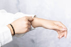 A close-up of a person's hands and forearms, showing one hand adjusting the cuff of a white long-sleeve shirt while wearing a black beaded bracelet with gold accents on the wrist.